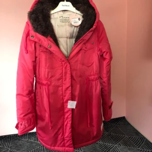 Röd parkajacka med foder - Snygg röd parkajacka från Ted Nicol med mysigt foder och huva. Jackan har dragkedja och knappar framtill samt justerbar midja för perfekt passform. Perfekt för kyliga dagar!