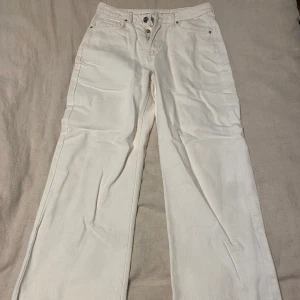 Vita jeans - Säljer ett par vita byxor i bra skick från Bik Bok. De har en normal passform och är högmidjade, vilket ger en snygg siluett. Perfekta för både vardag och festliga tillfällen. Byxorna har en klassisk design med knappar och dragkedja framtill.