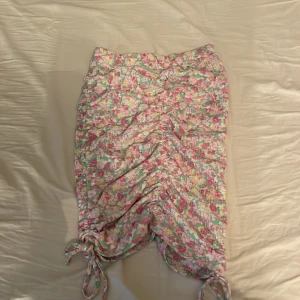 Blommig kjol från Gina Tricot - Söt blommig kjol från Gina Tricot i återvunnen polyester. Kjolen har ett färgglatt mönster i rosa, grönt och vitt med snörning på sidorna för en justerbar passform. Perfekt för en somrig look!