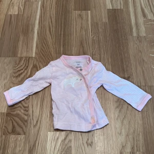 Rosa randig babytröja från Just One You - Söt rosa randig babytröja från Just One You med ett litet broderat djurmotiv på framsidan. Tröjan har långa ärmar och praktiska tryckknappar för enkel påklädning. Perfekt för de minsta!