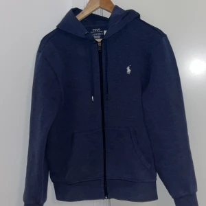 Blå hoodie från Polo Ralph Lauren - Snygg blå hoodie från Polo Ralph Lauren med dragkedja och klassisk logga på bröstet. Tröjan har en justerbar huva med snören och praktiska fickor framtill. Perfekt för en avslappnad stil.