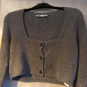 Grå croppad kofta från Zara - Säljer en stilren grå kofta från Zara. Den är croppad med tre knappar framtill och har långa ärmar. Perfekt för lager-på-lager under höst och vår.
