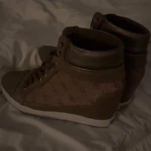 Guess skor med kilklack - Snygga bruna sneakers med glittriga detaljer och snörning. Skorna har en högre modell med en vit sula och ett mönster på sidan. De är en klack på ca 8-10cm. Skriv om ni har några frågor, hör gärna av er innan ni köper så aktiverar jag ”köp nu”💓