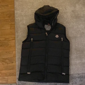 Svart dunväst från Moncler - Snygg svart dunväst från Moncler med huva och dragkedja. Västen har ett stilrent märke på bröstet och praktiska fickor med dragkedjor. Perfekt för kyligare dagar.