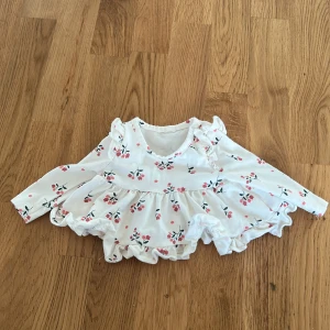 Vit blommig blus för barn - Söt vit blus med blommönster i rosa och grönt. Blusen har långa ärmar och volangdetaljer, perfekt för små barn. Tillverkad i mjuk bomull med lite stretch för bekvämlighet.