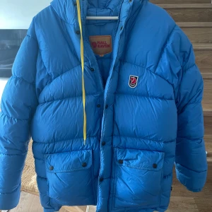 Blå dunjacka från Fjällräven - Säljer en blå dunjacka från Fjällräven med huva och praktiska fickor. Jackan har en dragkedja och knappar framtill samt en liten logotyp på bröstet. Perfekt för kalla dagar. Obs reva på höger ärm där av prisert. Behövs en tvätt!!