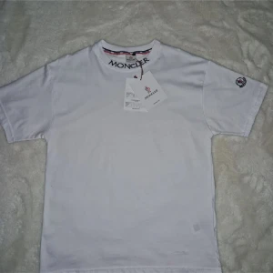Vit t-shirt från Moncler - Säljer en stilren vit t-shirt från Moncler med korta ärmar och broderad logga på ärmen. T-shirten har en klassisk rund halsringning och märkesdetaljer vid kragen. Perfekt för en avslappnad och trendig look.