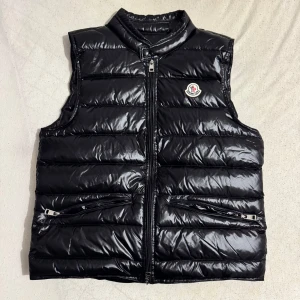 Svart dunväst från Moncler - Säljer en snygg svart dunväst från Moncler i mycket bra skick. Västen har en glansig finish och dragkedja framtill. Perfekt för höst och vår, med två praktiska fickor med dragkedja. Den är både stilren och funktionell, idealisk för lager-på-lager-stil.