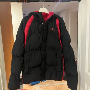 Svart och röd dunjacka från Jordan - Säljer en snygg och varm dunjacka från Jordan i svart med röda detaljer. Jackan har en pufferstil och är perfekt för vintern. Den har en dragkedja och långa ärmar, vilket gör den både praktisk och stilren. Perfekt för kalla dagar! 🖤❤️