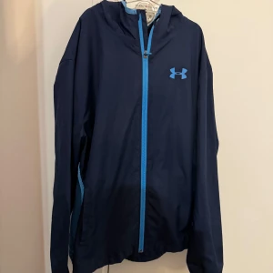 Mörkblå vindjacka från Under Armour 158/164 - Säljer en mörkblå vindjacka från Under Armour med ljusblå detaljer och dragkedja. Jackan har en luva och meshpanel på baksidan för extra ventilation. Perfekt för träning eller som en lätt ytterjacka.