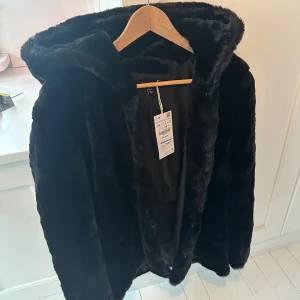 Svart fuskpälsjacka med huva från Zara - Säljer en svart fuskpälsjacka med huva från Zara. Jackan har en mjuk och fluffig känsla, perfekt för kyliga dagar. Den är långärmad och har en stilren design som passar till många olika outfits.                Den är mer som en Medium i storlek om man vill ha den lite oversize.