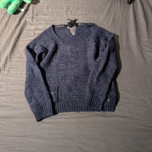 Mörkblå stickad tröja från Maison Scotch - Säljer en snygg mörkblå stickad tröja från Maison Scotch. Tröjan har en v-ringad design med en dekorativ knytning i nacken. Perfekt för kyliga dagar.