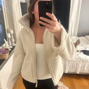 Beige teddyjacka med dragkedja - Mysig beige teddyjacka med dragkedja framtill. Perfekt för kyliga dagar med sin fluffiga och varma design. Jackan har en hög krage och långa ärmar, vilket ger extra värme och komfort.