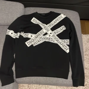 Svart tröja från Dsquared2 - Säljer en svart tröja från Dsquared2 i storlek M. Den har ett coolt korsmönster med Dsquared2-loggan i vitt över bröstet och ena ärmen. Perfekt för en stilren och modern look. Tröjan är i bra skick och passar både till vardags och fest.