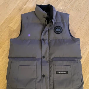 Grå dunväst från Canada Goose (black label) - Säljer en stilren grå dunväst från Canada Goose( black label) med broderad logga på bröstet. Västen har knappar och två stora fickor framtill. Perfekt för kyliga dagar.   