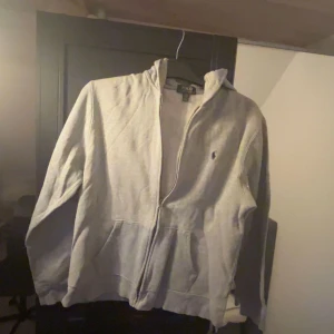Grå hoodie från Polo Ralph Lauren - Säljer en stilren grå hoodie från Polo Ralph Lauren med dragkedja och klassisk logga på bröstet.