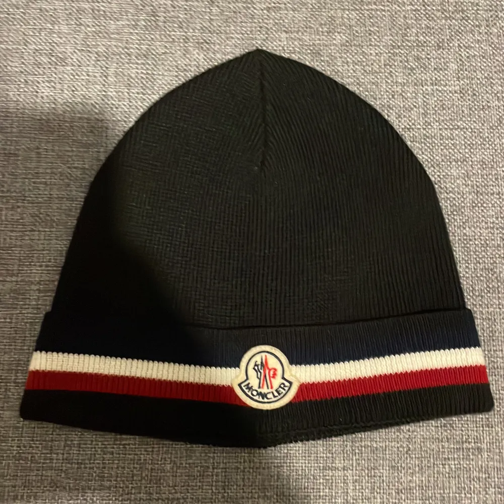 Snygg svart mössa från Moncler med ribbad design och klassisk logga framtill. Den har en stilren rand i vitt och rött runt kanten. Perfekt för att hålla värmen under kyliga dagar och samtidigt se trendig ut. Passar både till vardags och mer uppklädda tillfällen.. Accessoarer.