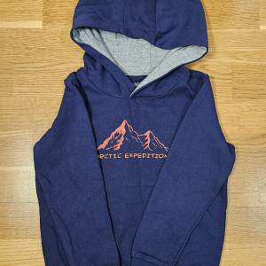 Säljer en snygg mörkblå hoodie med ett coolt bergstryck och texten 'Arctic Expedition' i orange. Perfekt för kalla dagar med sin mjuka insida och praktiska huva. Passar både till vardags och äventyr! 🏔️