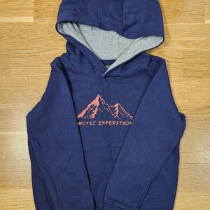 Mörkblå hoodie med tryck - Säljer en snygg mörkblå hoodie med ett coolt bergstryck och texten 'Arctic Expedition' i orange. Perfekt för kalla dagar med sin mjuka insida och praktiska huva. Passar både till vardags och äventyr! 🏔️