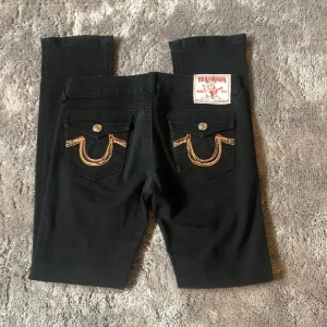 Svarta jeans från True Religion - Snygga svarta jeans från True Religion med ikonisk broderad detalj på bakfickorna. Jeansen har en klassisk femficksdesign och knappar i metall. Perfekta för en stilren look. Har endast används ett fåtal gånger av en tidigare ägare, då jag såg att de var förstora flr mig i midjan. Vid intresse, kontakta för fler bilder, pris kan diskuteras 