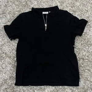 Calvin Klein T-Shirt - Snygg svart pikétröja från Calvin Klein med dragkedja framtill och klassisk krage. Perfekt för en stilren look.