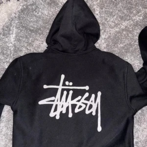 basic zip hopdie - Snygg svart hoodie från Stüssy med dragkedja och huva. Den har ett stort vitt Stüssy-tryck på ryggen och ett mindre på bröstet. Perfekt för en avslappnad stil.