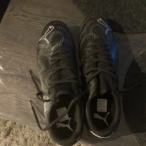 Svarta fotbollsskor från Puma - Snygga svarta fotbollsskor från Puma med vita detaljer och snörning. Skorna har dobbar för bra grepp på planen och en stilren design med Puma-loggan på sidan.