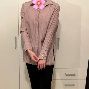 Lila/rosa långärmad blus - Säljer en elegant lila/rosa blus med långa ärmar och knappar vid ärmsluten. Blusen har en lös passform och är perfekt för både vardag och fest. Materialet ser ut att vara lätt och bekvämt, vilket gör den idealisk för vår och höst. Den har en klassisk krage och en stilren design.
