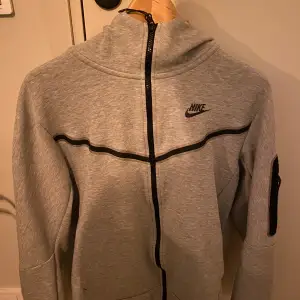 Säljer en grå hoodie från Nike i mycket bra skick. Den har en dragkedja framtill och en snygg svart detalj som går över bröstet. Perfekt för höst och vår, med långa ärmar och en bekväm passform. Idealisk för träning eller vardagsbruk. Pris kan diskuteras 