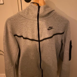Grå hoodie från Nike - Säljer en grå hoodie från Nike i mycket bra skick. Den har en dragkedja framtill och en snygg svart detalj som går över bröstet. Perfekt för höst och vår, med långa ärmar och en bekväm passform. Idealisk för träning eller vardagsbruk. Pris kan diskuteras 