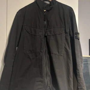 Svart jacka från Stone Island - Säljer en svart jacka från Stone Island i bra skick. Den har en dragkedja framtill och en broderad logga på ärmen. Perfekt för höst och vår med sin stilrena design. Jackan har långa ärmar och är gjord i ett slitstarkt material.