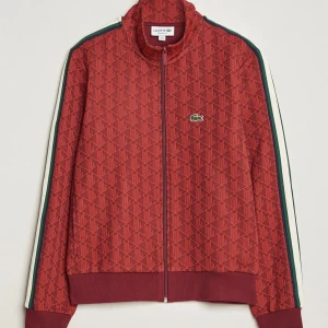 Tracksuit från Lacoste, som ny! - Snygg röd jacka från Lacoste med ett geometriskt mönster och klassisk krokodil-logga på bröstet. Jackan har dragkedja och ribbade muddar. Perfekt för en sportig look. Som ny, inga defekter. Julklappstips! Storlek S herr nypris 2000kr