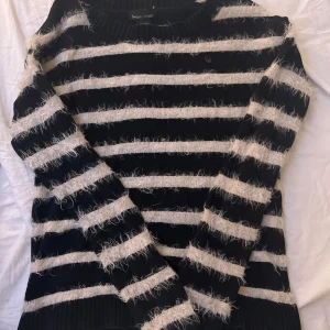 Svart och vit randig tröja från Sweater Project - Säljer en mysig svart och vit randig tröja från Sweater Project. Tröjan har en fluffig textur med långa ärmar och ribbstickade kanter. Perfekt för kyliga dagar!