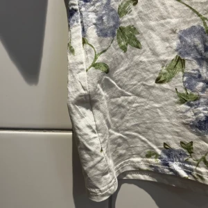Vit blommig t-shirt - Säljer en söt vit t-shirt med blå blommor och gröna blad. Den har en normal passform och korta ärmar, perfekt för vår och sommar. Materialet känns mjukt och bekvämt, idealiskt för en avslappnad stil. Passar både till jeans och kjol för en fräsch look!