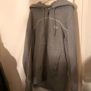 Grå hoodie från Hugo Boss - Säljer en stilren grå hoodie från Hugo Boss. Den har en dragkedja framtill och justerbar huva med snören. Perfekt för både vardag och avslappnade tillfällen. Den är i bra skick och redo för en ny ägare!