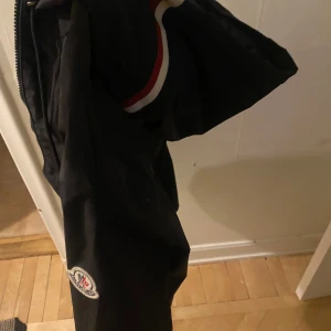 Svart jacka från Moncler - Snygg svart jacka från Moncler med dragkedja och huva. Jackan har Moncler-logga på ärmen och vita samt röda detaljer på huvan. Perfekt för en stilren look.