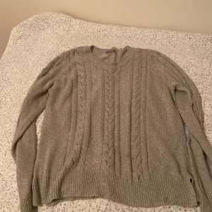 Grå stickad tröja från Lindex - Säljer en mysig grå stickad tröja från Lindex med flätmönster. Tröjan har långa ärmar och ribbstickade muddar vid ärmslut och nederkant. Perfekt för kyliga dagar!