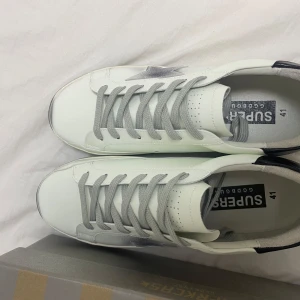 Golden Goose skor  - Snygga vita sneakers från Golden Goose med grå stjärndetalj och grå snörning. Skorna har en svart häl och en sliten look på sulan för en trendig stil. Perfekta för en avslappnad och stilren look. Helt ny och inga defekter! Hör av er vid frågor!!!