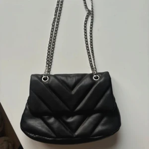 Svart quiltad axelväska med kedja - Svart axelväska i quiltat mönster med silverfärgad kedja som axelrem. Aldrig använd.
