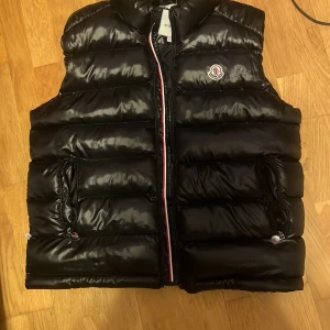 Svart dunväst från Moncler - Säljer en snygg svart dunväst från Moncler med dragkedja och broderad logga på bröstet. Västen har en stilren design med två praktiska fickor framtill. Perfekt för kyligare dagar.