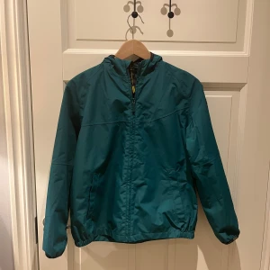 Grön vindjacka från Barbour - Snygg grön vindjacka från Barbour med dragkedja och huva. Jackan har långa ärmar och en stilren design, perfekt för blåsiga dagar. Den har en diskret logga på ärmen och elastiska muddar för en bekväm passform. Jackan är stl xl i barn passar även vuxen xs. 