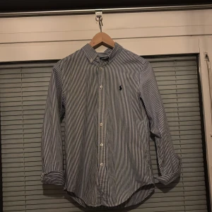 Randig skjorta från Ralph Lauren - Snygg blå- och vitrandig skjorta från Ralph Lauren med klassisk krage och knappar framtill. Skjortan har ett broderat märke på bröstet och långa ärmar. Perfekt för en stilren look det är storlek 14y skriv om ni har några funderingar! Skick 8/10