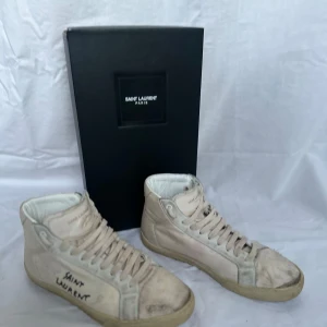 Vita sneakers från Saint Laurent - Snygga vita sneakers från Saint Laurent med hög design och klassisk snörning. Skorna har en stilren look med märkets logga på sidan. Perfekta för en trendig och avslappnad stil. Storlek 42 lite smutsiga men inget fel med dom OBS inget og medföljer