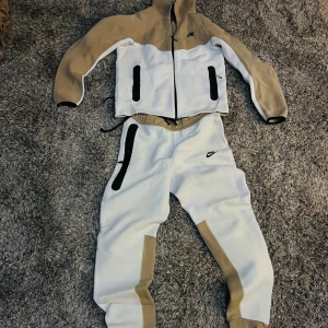 Nike Tech - Snygg träningsdräkt från Nike i beige och vitt. Jackan har dragkedja och svarta detaljer, medan byxorna har elastisk midja och dragkedjor vid fickorna. Perfekt för en sportig look.