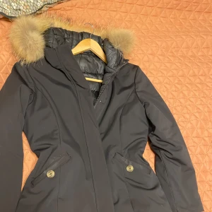 Svart parkajacka med pälsdetalj - Snygg svart parkajacka med avtagbar pälsdetalj på huvan. Jackan har dragkedja och knappar framtill samt två fickor med knappar. Perfekt för kyliga dagar. Pälsen är äkta och pris kan såklart diskuteras. Passar m och l