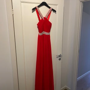 Röd långklänning med pärldetaljer - Elegant röd långklänning med pärlbeströdda axelband och midja. Klänningen har en vacker drapering framtill och en smickrande passform. Perfekt för speciella tillfällen.