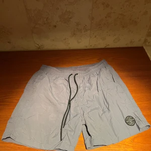 Grå badshorts från Surfline - Snygga grå badshorts från Surfline med en cool patch på benet. De har en elastisk midja med snörning för perfekt passform.