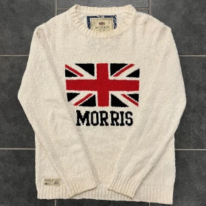 Vit stickad tröja från Morris - Säljer en vit stickad tröja från Morris med ett stort Union Jack-motiv och texten 'Morris' på framsidan. Tröjan är tillverkad i 100% bomull och har långa ärmar samt ribbade kanter. Perfekt för en stilren och bekväm look.