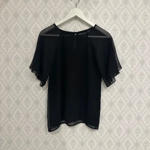 Svart transparent blus - Snygg svart transparent blus med korta ärmar och rund hals. Blusen har en elegant design med en liten knappdetalj i nacken. Perfekt för att lägga till en touch av stil till din garderob.