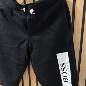 Svarta shorts från Hugo Boss - Snygga svarta shorts från Hugo Boss med vit logga på sidan. De har en bekväm passform och dragsko i midjan för justerbarhet. Perfekta för en avslappnad stil. Storlek 14/162 cm från kidsbrandstore. Är använda men i bra skick! 